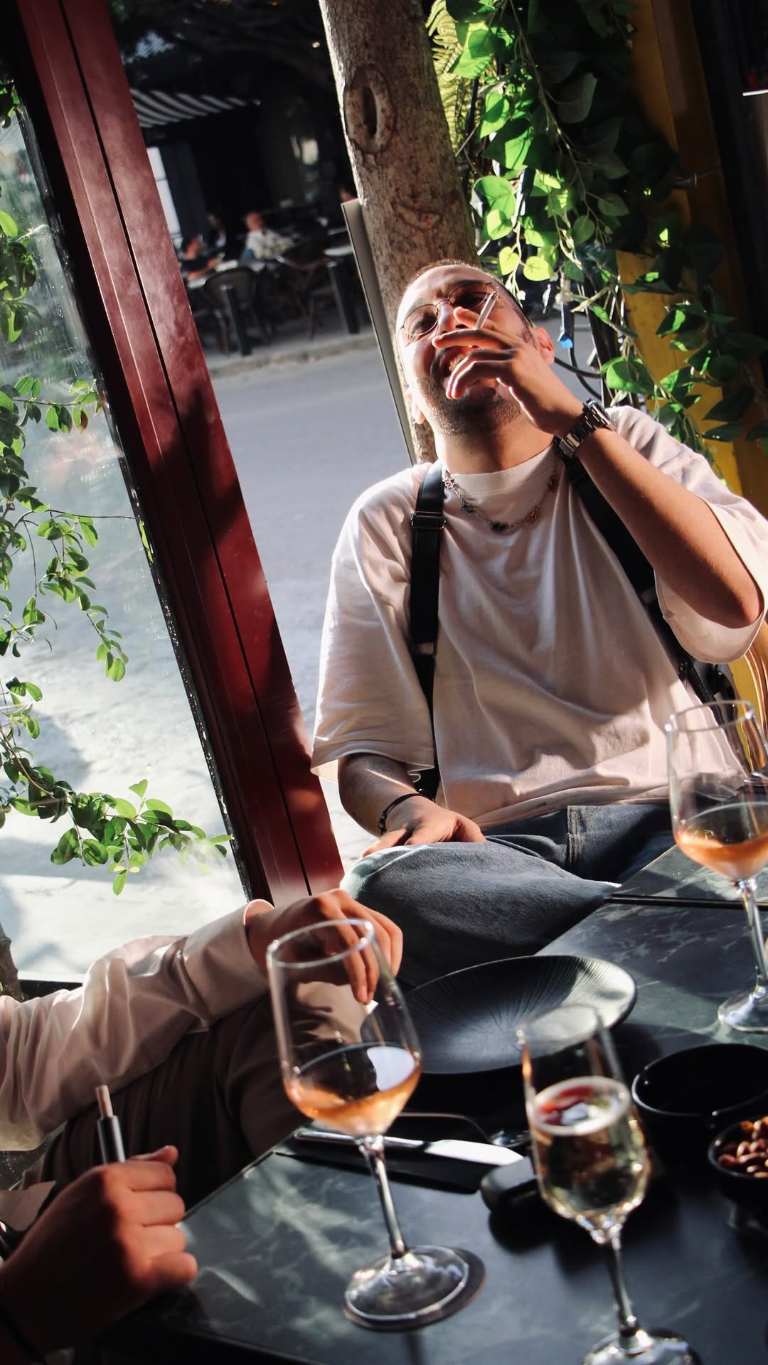A guest laughing over wine glasses on the Maia terrace, sunlight filtering through lush greenery