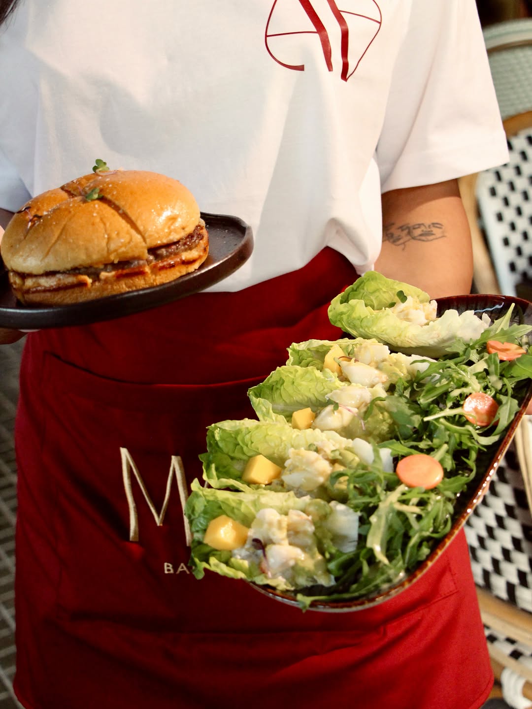 A Maia staff member in a red apron, the MAIA wordmark visible, carrying a Wagyu burger and a salad
