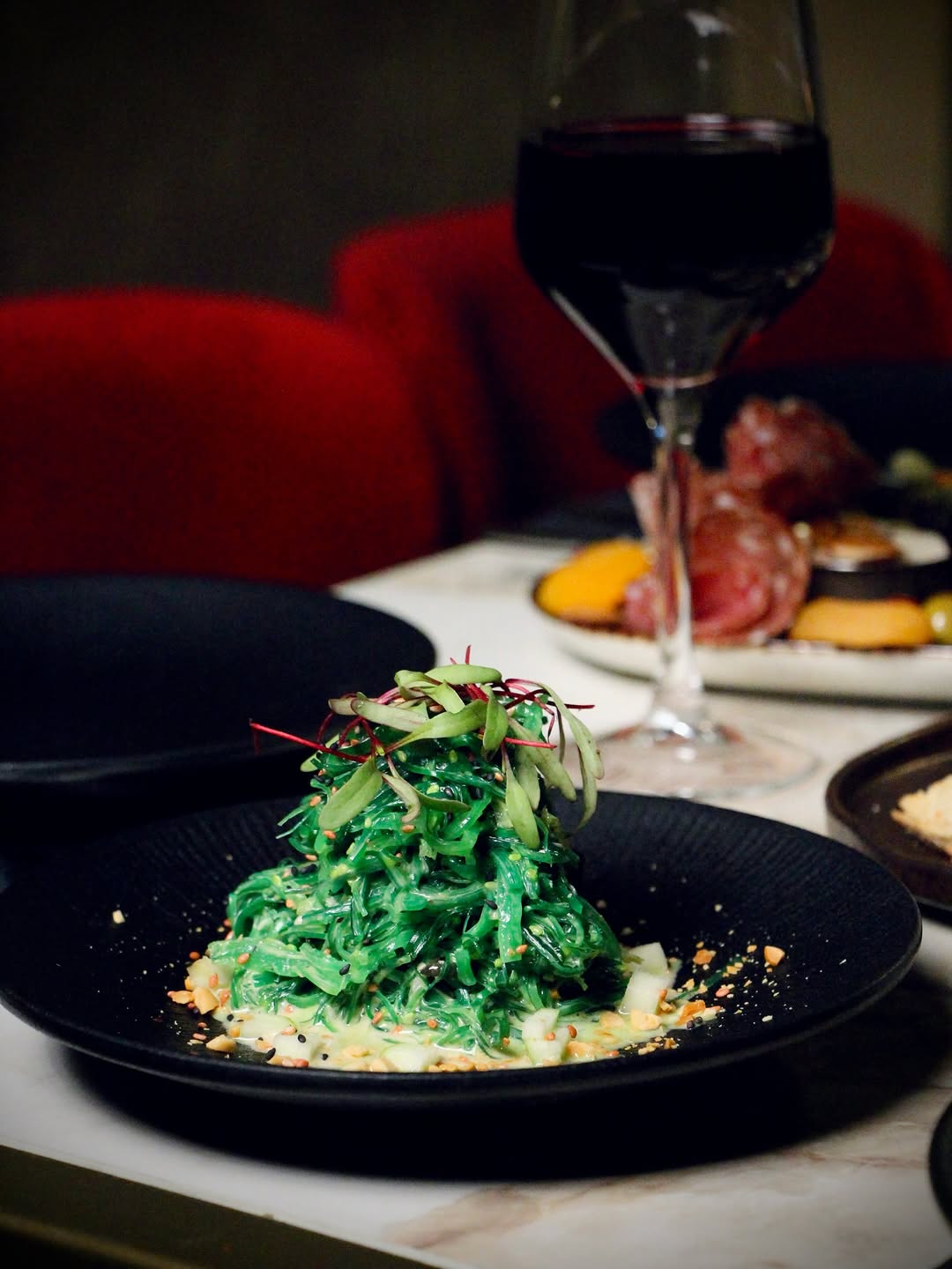 A vivid green wakame seaweed salad mounded high on a black plate, red wine and steak in the background