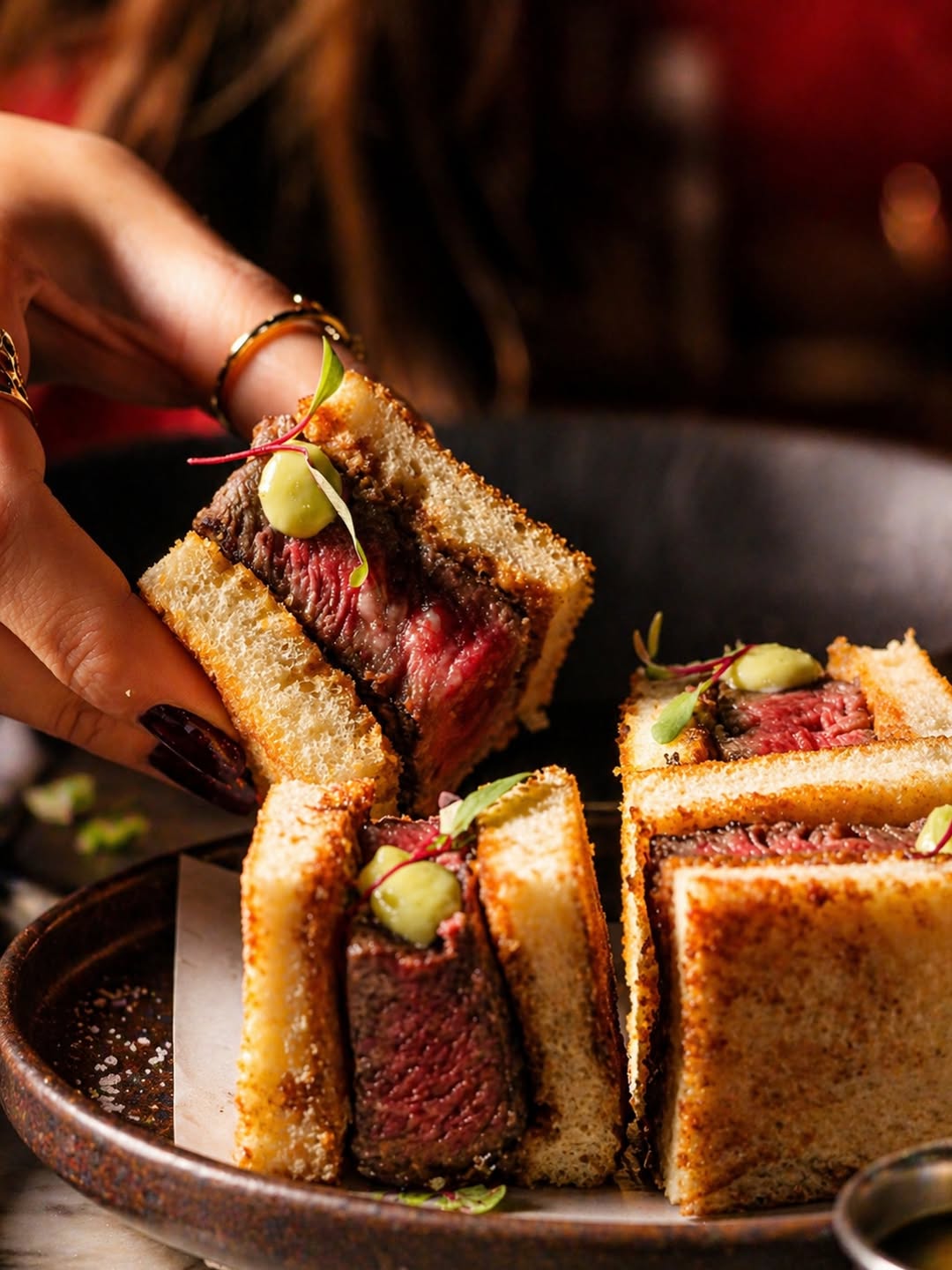 Medium-rare A5 Wagyu sando between toasted Japanese milk bread, lifted by a jewel-ringed hand